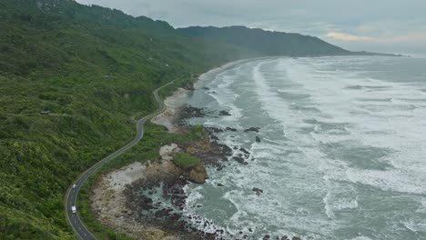 Aerial-flight-following-a-tourist-campervan-driving-on-a-wild,-rugged-and-remote-coastal-road-on-the-West-Coast,-South-Island-of-New-Zealand-Aotearoa