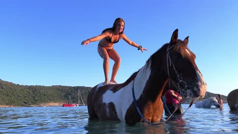 Little-girl-standing-balancing-on-the-back-of-a-horse-immersed-in-sea-water-in-summer-season