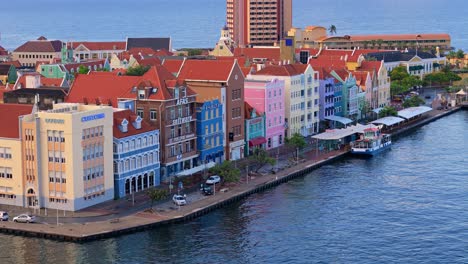 Cars-drive-below-colorful-buildings-of-Willemstad-Curacao-at-sunrise,-Handelskade,-Punda-District