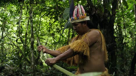 An-indigenous-guy-wearing-a-feathered-hat-and-fringed-shirt-peels-young-coconut-leaves-in-Leticia,-Colombia