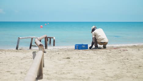A-black-male,-wearing-a-baseball-cap,-diligently-cleans-the-beach-with-hands-and-a-rake,-against-a-backdrop-of-crystal-blue-water