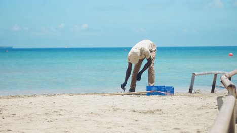 Un-Hombre-Negro,-Con-Una-Gorra-De-Béisbol,-Limpia-Diligentemente-La-Playa-Con-Las-Manos-Y-Un-Rastrillo,-Recogiendo-Basura-Y-Colocándola-En-Una-Caja-De-Pescado-Azul