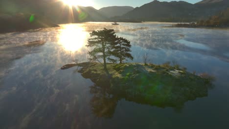 Small-island-in-calm-lake-with-gentle-mist-curling-on-water-surface-with-orbit-revealing-rising-sun-flare-and-reflection-occluded-by-trees