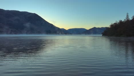 Flying-low-over-calm-lake-surface-as-mist-gently-moves-along-prior-to-dawn-in-autumn