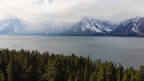 A-low-flying,-orbital-drone-shot-along-the-forest-covered-coastline-of-Jackson-Lake,-with-the-Grand-Teton-Range-in-the-background,-in-Grand-Teton-National-Park-of-Northwestern-Wyoming