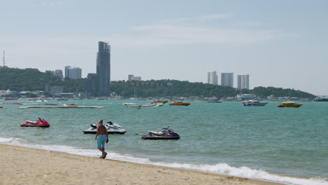 Foreign-tourists-taking-a-stroll-at-the-sandy-beachfront-of-Pattaya-Beach,-against-the-background-of-the-business-hub-of-Chonburi-province-in-Thailand