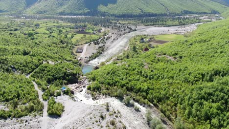 Drohnenansicht-In-Albanien-In-Den-Alpen,-Die-über-Einen-Kristallklaren-Fluss-Mit-Felsigem-Boden-Und-Grünem-Wald-An-Den-Seiten-Im-Theth-Fliegt