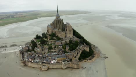 Cámara-Drone-Mont-Saint-Michel-Mirando-Hacia-Atrás,-Hay-Muchos-Turistas-Que-Regresan-Después-De-Ver-El-Castillo