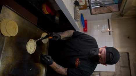 Vertical-slow-motion-of-a-latin-hispanic-man-dressed-in-black-with-latex-gloves-and-a-mask-preparing-a-quesadilla-on-a-griddle-using-a-steel-spatula