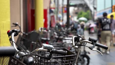 Nahaufnahme-Der-Geparkten-Fahrräder-Im-Shinsekai-Viertel-An-Einem-Regnerischen-Tag-Mit-Menschen,-Die-Im-Hintergrund-Vorbeigehen