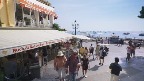 Amalfi-Positano-Italia-Viajes-De-Inmersión-Turismo-Mar-Mediterráneo-Costa-Agua-Europa,-Caminar,-4k-|-Hermosa-Mujer-Con-Tatuajes-Pasando-Cerca-De-Una-Icónica-Plaza-Llena-De-Gente.
