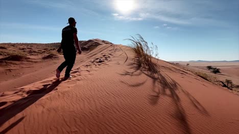 Man-wearing-a-hat-walks-over-a-red-sand-dune-in-the-desert-of-Namibia-with-the-sun-casting-beautiful-shadows-of-the-man-and-some-grass-blowing-in-the-wind