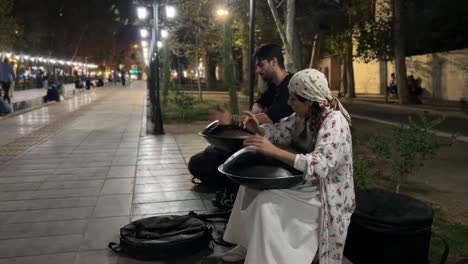 Street-Music-performance-in-the-big-city-at-night-a-couple-playing-hang-guitar-in-pavement-city-center-downtown-dark-vibe-happy-song-park-light-people-life-Tehran-Iran-walking-promenade-artist-woman