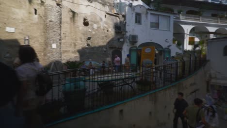 Amalfi-Positano-Italy-Immersive-Travel-Tourism-Mediterranean-Sea-Coast-Water-Europe,-Walking,-4K-|-Moving-Down-Stairs-Through-Busy-Tourist-Crowd-in-Shadows-Underneath-Mountainside-Dwellings-Above
