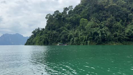 Khao-Sok-Lake-in-Thailand:-A-boat-gently-gliding-along-the-lush,-vegetation-covered-shoreline,-creating-a-tranquil-scene-on-the-water