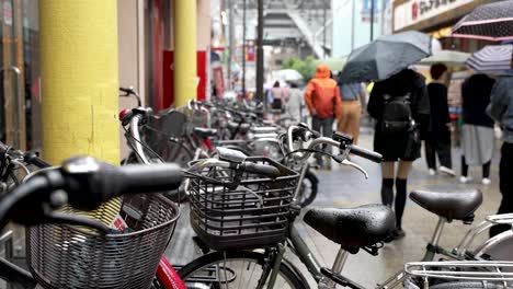 Reihe-Geparkter-Fahrräder-Im-Shinsekai-Viertel-An-Einem-Regnerischen-Tag,-Im-Hintergrund-Laufen-Menschen-Vorbei