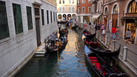 Venetian-canal-full-of-gondolas-with-passengers,-gondoliers-and-tourists-walking-along-sidewalk-in-Venice,-Italy