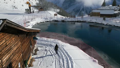 Tourist-walking-by-a-winter-log-cabin-located-near-the-mountainside-in-Engelberg,-Brunni,-Bahnen-in-Switzerland