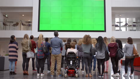 Students-applaud-a-big-screen-in-university-foyer,-back-view