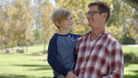 Father-carrying-seven-year-old-son-smiles-to-camera-in-park
