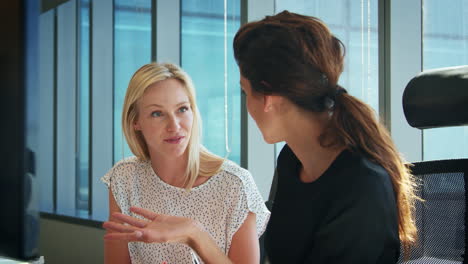 Two-Businesswomen-Working-At-Desks-Have-Discussion-Together