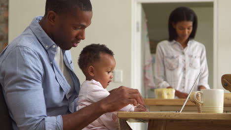 Father-And-Baby-Daughter-Use-Laptop-As-Mother-Prepares-Meal
