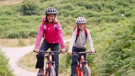 Two-children-having-fun-on-mountain-bikes-in-the-countryside-ride-past-the-camera-and-out-of-shot,-Lake-District,-UK