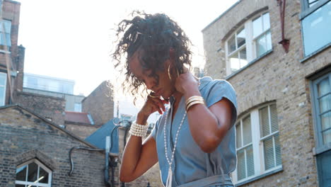 Fashionable-young-black-woman-standing-in-front-of-old-buildings-adjusting-her-necklace,-lens-flare,-low-angle