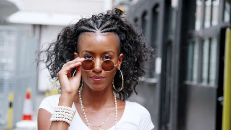 Moda-Joven-Mujer-Negra-Girando-Y-Mirando-A-La-Cámara-Sobre-Sus-Gafas-De-Sol-Redondas,-Cerrar