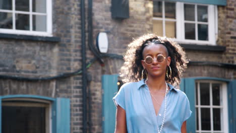 Fashionable-young-black-woman-wearing-blue-dress-and-sunglasses-walking-past-camera-in-the-street,-backlit,-low-angle