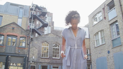 Fashionable-young-black-woman-in-blue-dress-standing-in-the-sun-against-buildings,-lens-flare,-low-angle