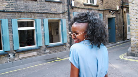 Trendy-young-black-woman-in-blue-dress-and-sunglasses-walking-on-the-street-laughing,-back-view,-close-up