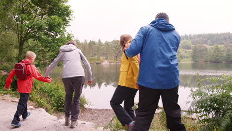 Familia-Joven-Caminando-Hacia-Un-Lago-Y-Jugando-En-La-Orilla,-Vista-Posterior,-Lake-District,-Reino-Unido