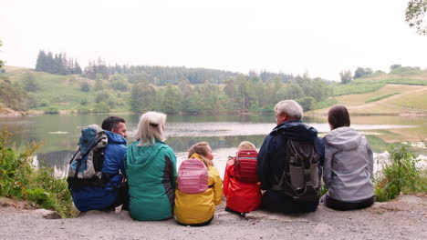 Multi-generation-family-sitting-together-in-a-row-talking-by-a-lake,-back-view,-Lake-District,-UK