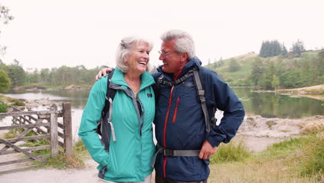 Senior-couple-on-a-camping-holiday-standing-by-a-lake-laughing,-close-up,-Lake-District,-UK