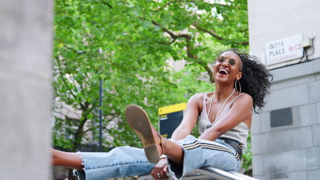 Trendy-young-black-woman-in-camisole-and-side-stripe-jeans-has-fun-sitting-on-a-handrail-in-a-city-street,-low-angle
