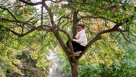 Mixed-race-girl-sitting-in-an-Autumn-tree,-smiling-to-camera,-low-angle