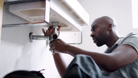 Young-black-male-plumber-sitting-on-the-floor-replacing-the-trap-pipe-under-a-bathroom-sink,-low-angle