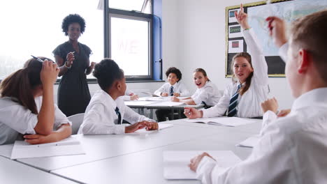 Female-High-School-Tutor-Asking-Pupils-Wearing-Uniform-Sitting-At-Desks-Question-To-Which-They-Raise-Their-Hands