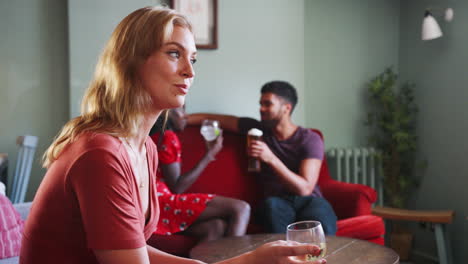 Close-up-side-view-of-young-blonde-woman-sitting-and-talking-with-her-friend-in-the-lounge-room-at-a-pub,-focus-on-foreground