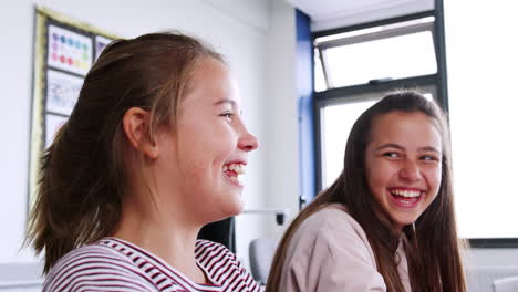 Two-Female-High-School-Students-Laughing-As-They-Sit-In-Classroom