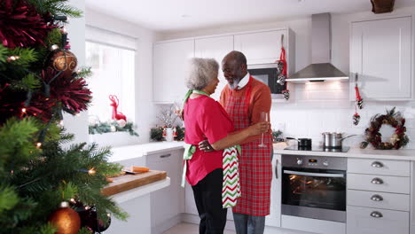 Happy-mature-black-couple-holding-champagne-glasses,-laughing-and-dancing-in-the-kitchen-while-preparing-Christmas-dinner,-side-view