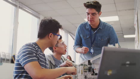 Teacher-With-Two-Male-College-Students-Building-Machine-In-Science-Robotics-Or-Engineering-Class