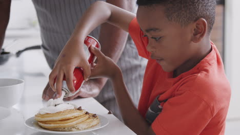 Grandfather-In-Kitchen-With-Grandson-Eating-Pancakes-Together