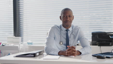 Portrait-Of-Smiling-Male-Doctor-Sitting-Behind-Desk-In-Office