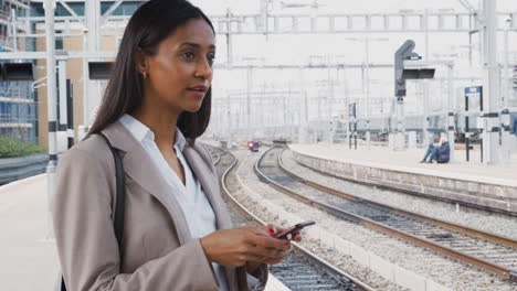 Businesswoman-Commuting-To-Work-Standing-On-Train-Platform-Using-Mobile-Phone