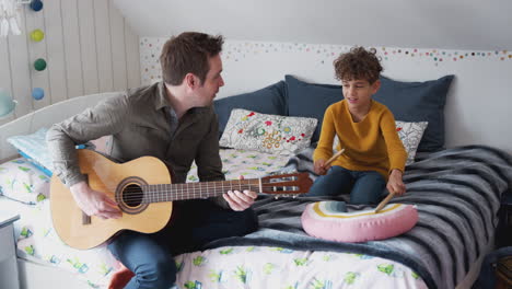 Alleinerziehender-Vater-Spielt-Gitarre-Mit-Sohn,-Der-Im-Schlafzimmer-Auf-Einem-Kissen-Trommelt