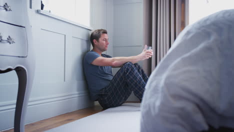 Depressed-Man-Wearing-Pajamas-Sitting-On-Floor-Of-Bedroom-Holding-Glass-Of-Whisky