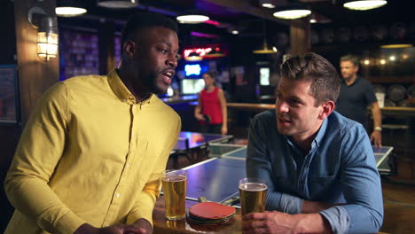 Male-Friends-Meeting-For-Drinks-In-Bar-With-Customers-Playing-Table-Tennis-Behind