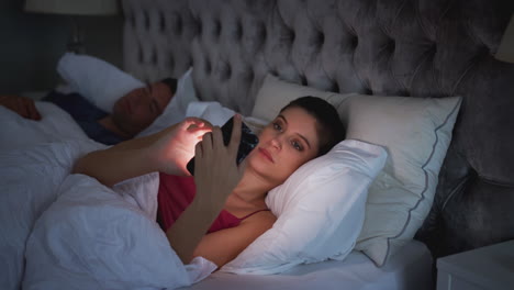 Frau-Liegt-Im-Bett-Und-Checkt-Ihr-Mobiltelefon,-Während-Ein-Mann-Neben-Ihr-Schläft